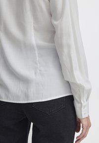 Camicia bianca a maniche lunghe con una consistenza liscia e sottili linee verticali. Presenta polsini con bottoni e un orlo curvo sopra jeans scuri.