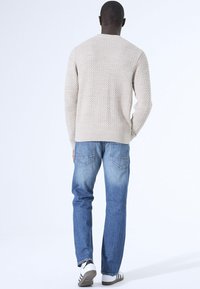 Beiger Strickpullover mit einem strukturierten Muster, kombiniert mit hellblauen Jeans mit Gesäßtaschen und weißen Sneakers mit dunklen Akzenten.