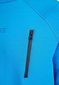 Suéter de forro polar azul con un bolsillo negro con cremallera en el pecho. Presenta una tela texturizada y costuras de contraste a lo largo de las costuras.