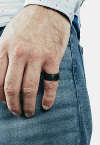 Schwarzer strukturierter Ring an einer Hand mit einer glatten schwarzen Einlage. Die Hand ruht auf einer Jeans, die den Hautton und die Haarstruktur zeigt.
