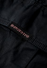 Zwarte stof met een getextureerde afwerking, voorzien van een zwarte label met roze tekst en logo "SUPERDRY". Eenvoudige, strakke gestikte afwerking.
