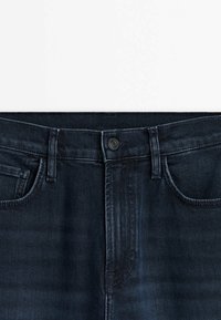 Gros plan sur la ceinture d’un jean en denim bleu foncé avec bouton, passants de ceinture et poches avant sur fond blanc.