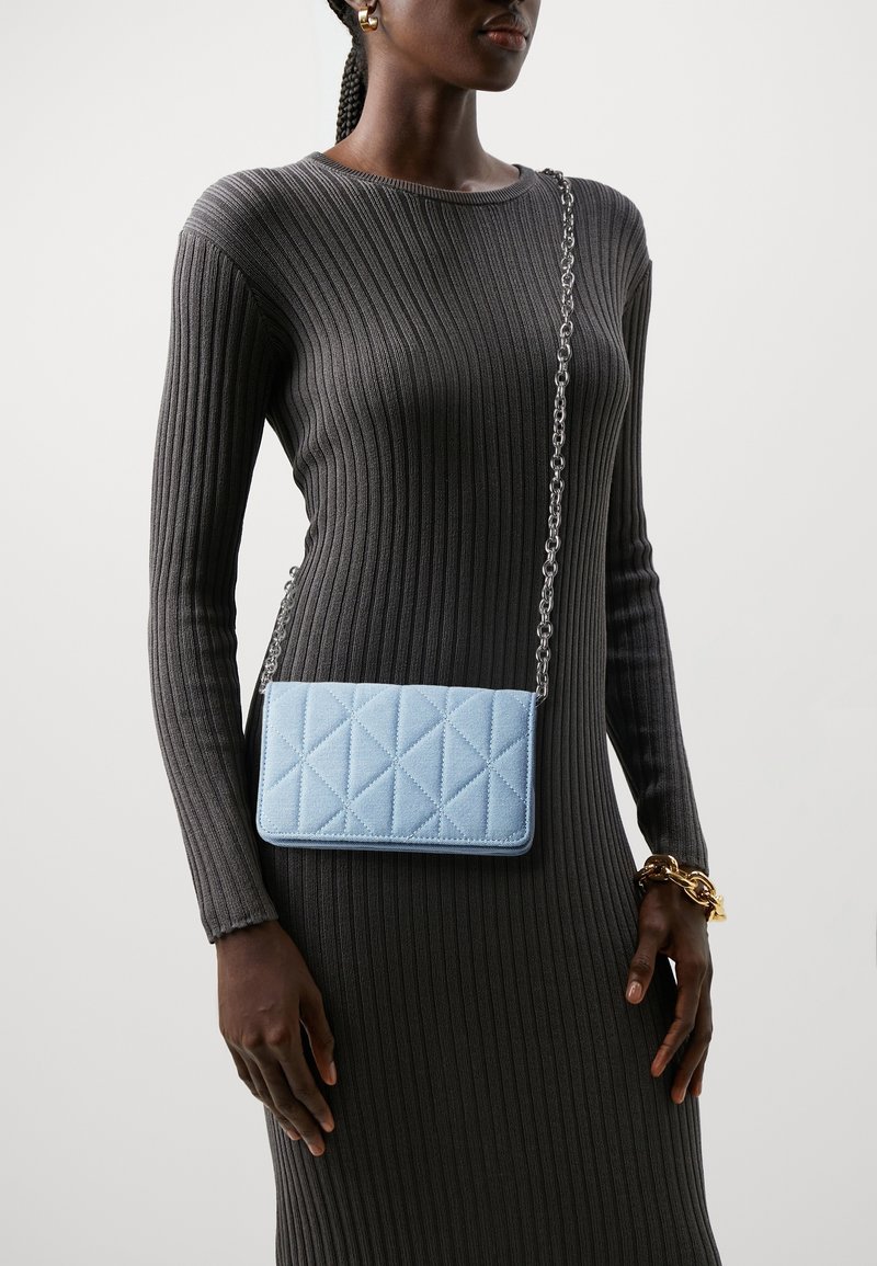 Bolso acolchado de color azul claro con una correa de cadena, forma rectangular, superficie texturizada, y acentuado con una cadena en tono plateado.