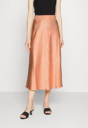 Femme portant une jupe midi pêche brillante avec un motif paisley subtil, un top noir sans manches et des sandales noires à talons ouverts.
