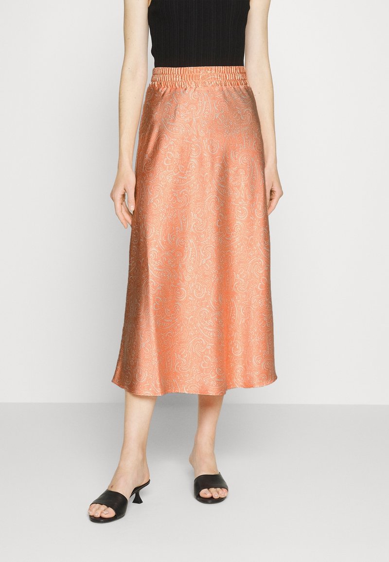 Femme portant une jupe midi pêche brillante avec un motif paisley subtil, un top noir sans manches et des sandales noires à talons ouverts.