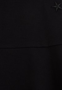 Prenda de tela negra con una textura suave, dos paneles horizontales distintos y un pequeño detalle de estrella bordado en la esquina superior.