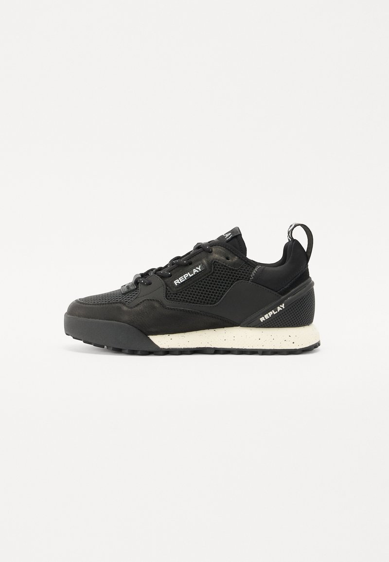 Zapatilla deportiva negra con superposiciones de malla y sintético, con una suela salpicada en blanco y un logotipo en el costado y en la lengüeta.