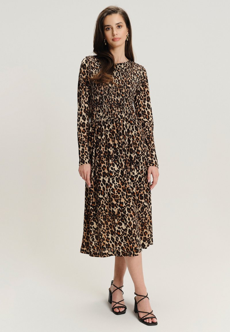 Middellange jurk met lange mouwen in luipaardprint, met een smock-borststuk, vloeiende rok en een effen crèmekleurige achtergrond. Zwarte sandalen met banden.