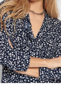 Blouse bleu marine avec imprimé floral blanc, dotée d'un col volanté et de manches longues. Le tissu a une texture lisse avec un léger éclat.
