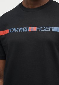 Μαύρο βαμβακερό μπλουζάκι με τολμηρό λογότυπο «TOMMY HILFIGER» σε μπλε και κόκκινο. Κοντά μανίκια και στρογγυλή λαιμόκοψη. Λεπτή υφή.