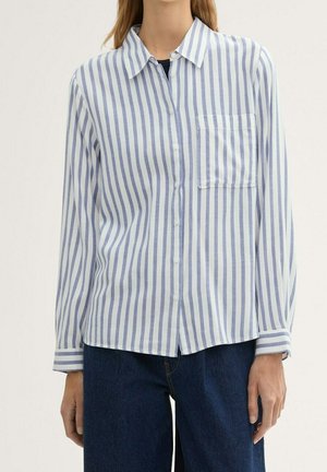 Vrouw die een wit overhemd met verticale blauwe strepen en een borstzak draagt, gecombineerd met donkerblauwe broek.