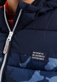 Nahaufnahme einer marineblauen und blauen Camouflage-Pufferjacke mit weißem Reißverschluss und einem Patch "Authentische Wildniserfahrung" auf der Brust.