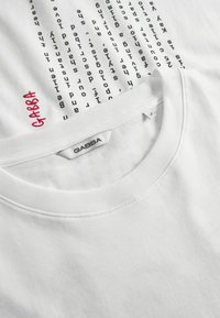 Tricou alb din bumbac cu un design vertical din text negru și logo-ul roșu "GABBA" pe piept. Guler ribatabil. Textură moale.