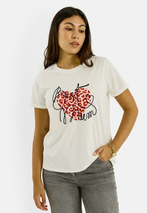 Femme portant un T-shirt blanc avec un cœur imprimé léopard rouge et un texte en cursive noire, associé à un jean gris et un bracelet.