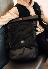 Person med beige hoodie och svart väst som håller en svart Johnny Urban ryggsäck med elastisk bungee-kabel och rullupplock.