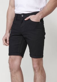 Mies, joka on pukeutunut käännettyihin mustiin farkkushortseihin ja valkoiseen t-paitaan, pitäen yhtä kättä shortsejensa taskussa yksinkertaista taustaa vasten.