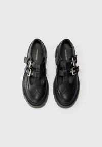 Chaussures en cuir noir avec deux sangles à boucle, ornées de détails brogues et dotées de semelles en caoutchouc épaisses. Texture lisse et forme arrondie.