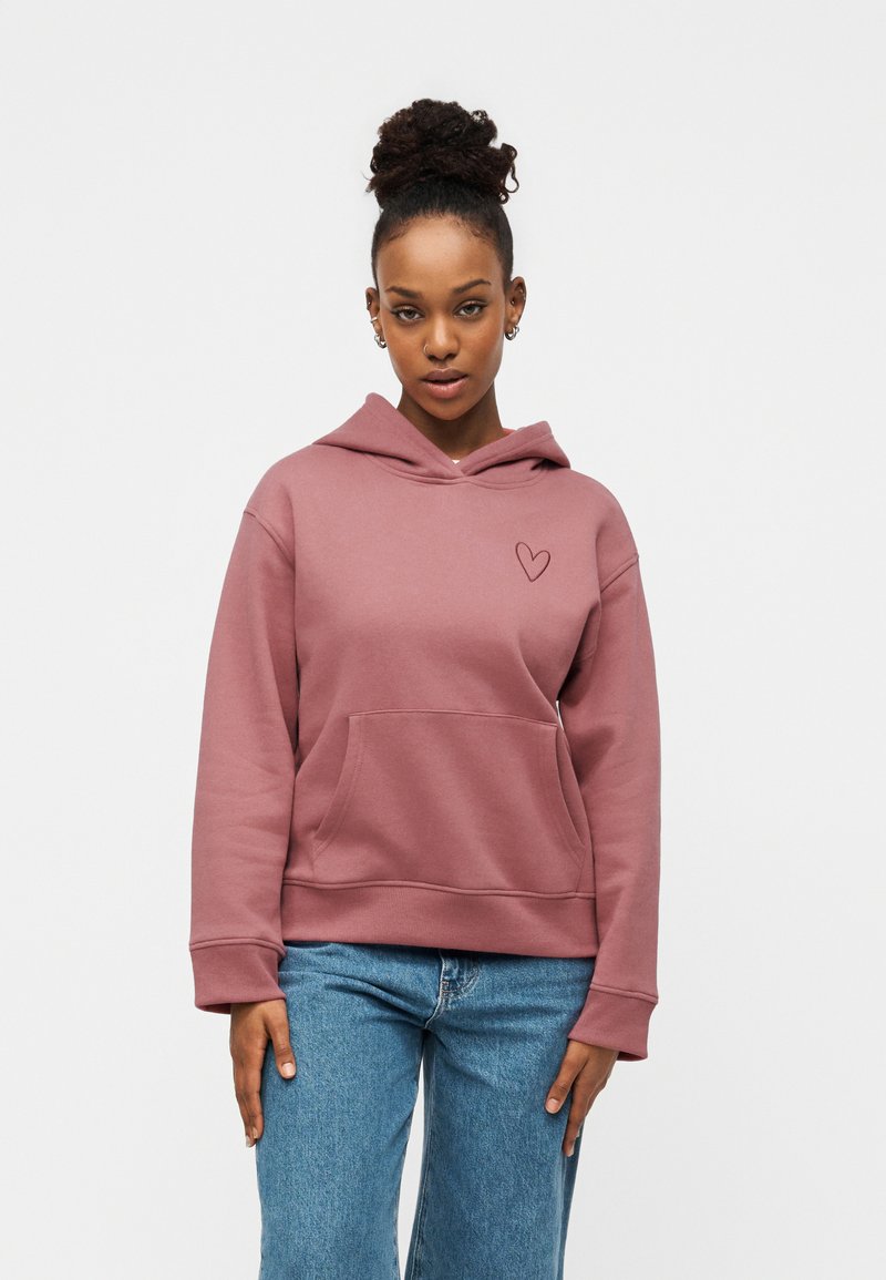 Frau mit lockigem Haar zu einem Dutt gebunden, trägt einen staubig rosa Hoodie mit einer kleinen Herzbestickung und blaue Jeans, steht vor einem weißen Hintergrund.