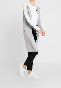 Cárdigan largo y gris con mangas blancas, hecho de un material de punto suave. Presenta bolsillos delanteros y botones negros, usado sobre una camisa blanca.