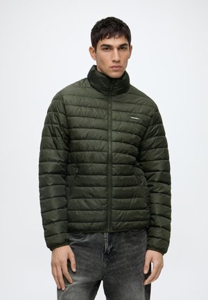 Jeune homme portant une veste matelassée vert foncé avec une fermeture éclair à l'avant et un col montant, associée à un jean noir délavé, debout devant un fond uni.