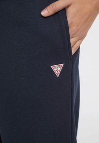 Navy blue cotton joggers with a fitted design featuring a small red and white triangular ‘GUESS’ logo on the left pocket.