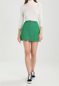 Minifalda de mezclilla verde con cremallera frontal, combinada con una blusa de manga larga blanca y zapatillas negras. Textura suave, diseño casual.