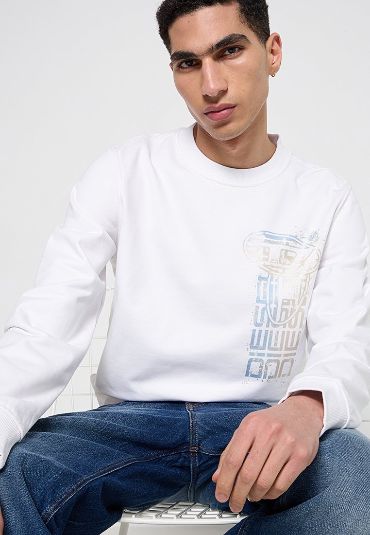 Felpa bianca con scollo rotondo, caratterizzata da una leggera stampa grafica blu e beige sul lato sinistro del petto. Abbinata a jeans blu scuro.