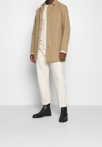 Světle béžový vnější kabát, zapnutý, vyrobený z látky s měkkou strukturou. Stylizován přes světelnou košili a bílé zkrácené kalhoty, doplněný černými botami.