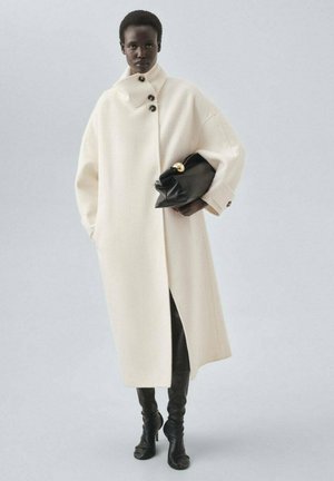 Übergroßer weißer Wollmantel mit hohem Kragen und drei schwarzen Knöpfen, kombiniert mit einer schwarzen Clutch und einer figurbetonten schwarzen Hose. Weiche Textur.