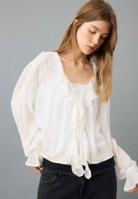 Bílá lehká halenka s volánkovým výstřihem a manžetami, doplněná středovým zavazováním. Dlouhé rukávy, měkká látka a volný střih.