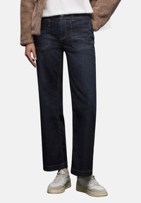 Granatowe dżinsy w szerokim kroju z kontrastowym pomarańczowym przeszyciem, pięcioma kieszeniami i zapięciem na guzik. Noszone z beżowymi trampkami.