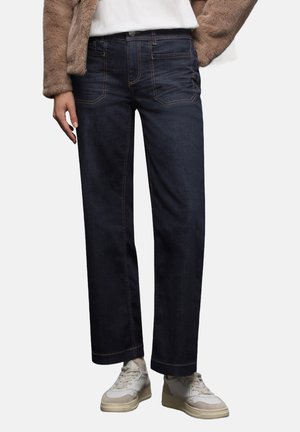 Dunkelblaue weite Jeans mit kontrastierender orangefarbener Naht, fünf Taschen und Knopfverschluss. Getragen mit beigen Sneakers.