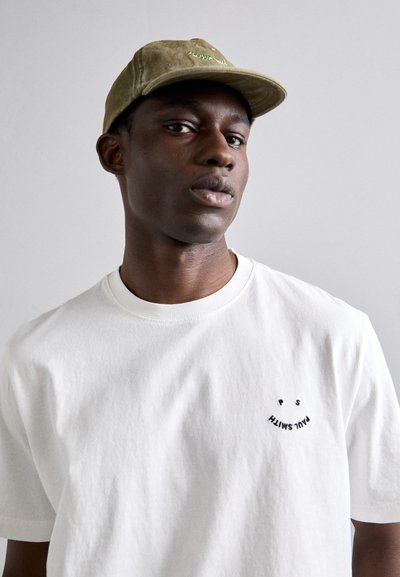 Λευκό βαμβακερό T-shirt με στρογγυλή λαιμόκοψη και κοντά μανίκια, που διαθέτει ένα μαύρο κεντημένο χαμόγελο και κείμενο στο αριστερό στήθος. Ελιές πράσινο καπέλο.