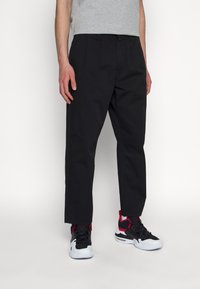 Homme portant un pantalon noir à jambes droites, un t-shirt gris et des baskets de sport noires, blanches et rouges, debout devant un fond blanc.