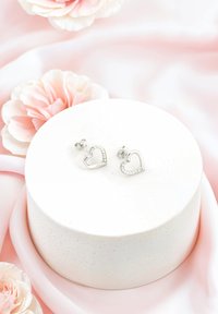 Söta hjärtformade silverörhängen med klara stenaccenter, visade på en strukturerad vit rund piedestal bland mjuka rosa tyger och blommor.