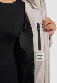 Intérieur d'une veste rose clair avec doublure noire, comportant une poche zippée et une étiquette en tissu qui indique "Un câlin chaud fait de tissu."