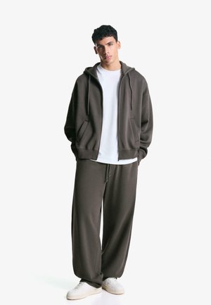 Brun zip-up hoodie med en hvid t-shirt under og matchende brune sweatpants. Stoffet ser blødt og afslappet ud. Hvide sneakers.