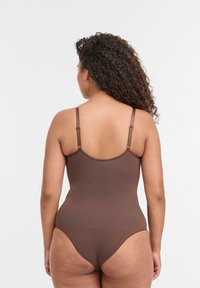 Donna con capelli ricci che indossa un bodysuit senza maniche di colore marrone scuro, rivolta di spalle su uno sfondo bianco semplice.
