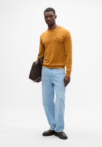 Musztardowy sweter, jasnoniebieskie dżinsy i ciemnobrązowa torba. Model stoi w luźnym kroju i gładkich fakturach. Prosty okrągły dekolt.