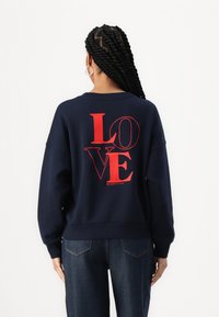 Námornícky modrý sveter s červenou grafikou "LOVE" potlačenou na chrbte, s okrúhlym výstrihom a rebrovanými rukávmi. Vyrobený z jemného materiálu.