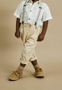 Beżowe lniane spodnie z podwiązanymi nogawkami w połączeniu z białą koszulą z krótkim rękawem i szarymi szelkami. Brązowe buty o teksturowanym wykończeniu.