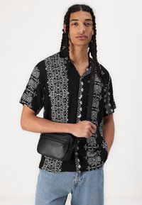 Cămașă neagră cu mânecă scurtă și model floral alb, design cu nasturi. Modelul ține o mică geantă neagră tip crossbody pe un fundal deschis.