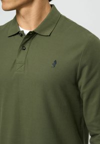 Oliva zöld hosszú ujjú pólóing, amely texturált anyagból készült, klasszikus gallérral és egy kis sötétkék logóval a mellkasán.