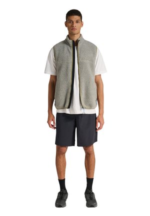 Jonge man met een lichtgrijze fleece bodywarmer over een wit T-shirt, zwarte korte broek, zwarte sokken en donkere sportschoenen, staand tegen een witte achtergrond.