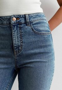 Blå denimjeans med hög midja, detaljer i kopparnitar och subtila förslitningar. Klassisk femficksdesign med kontrasterande sömmar.