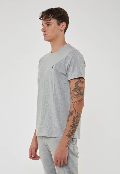 T-shirt in cotone grigio con maniche corte, collo a giro e piccolo logo sul petto. Il tessuto ha una consistenza liscia. Modello che indossa jeans grigio chiaro.