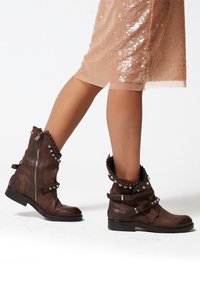 Brown leather ankle boots with decorative studs, zip detail, and layered cuff design. Flat sole with sturdy construction.