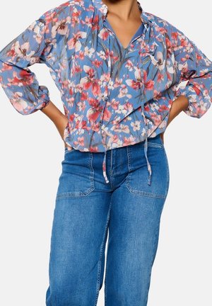 Kvinde iført en blå blomstret bluse med lange ærmer og opsamlede manchetter samt højtaljede blå jeans, hænderne hvilende på hoften.