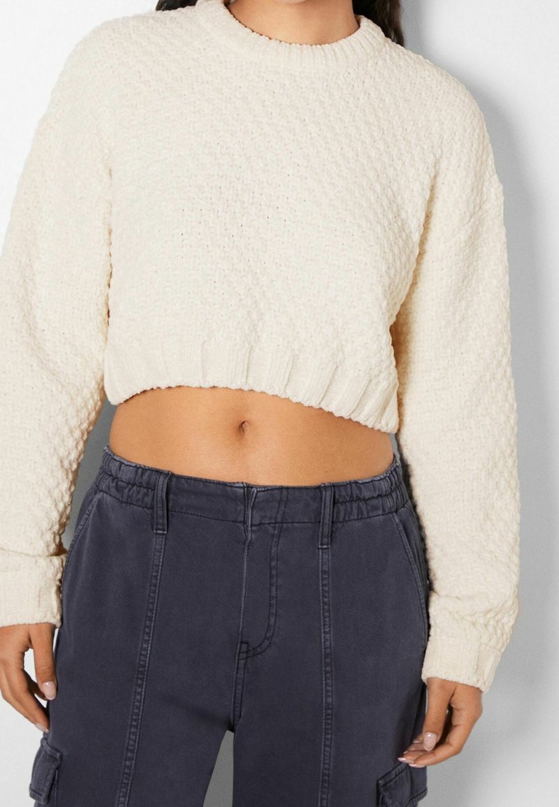 Personne portant un pull court crème texturé et un pantalon taille haute gris foncé avec ceinture élastique et poches latérales.