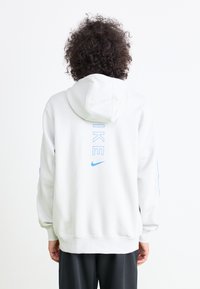 Sweatshirt branco com capuz, com a marcação azul da Nike nas costas e nas mangas, apresentando um corte descontraído e material suave.